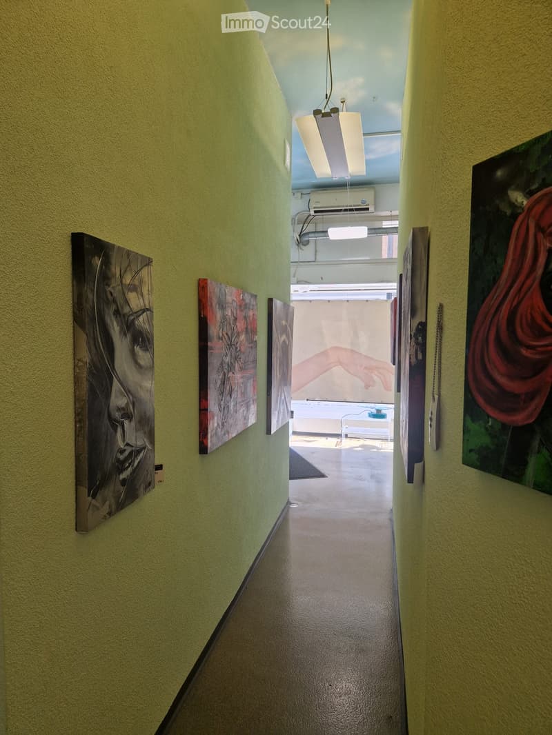 A narrow hallway with light green walls, several modern art pieces displayed on the walls, and a visible room at the end with a table and chairs.