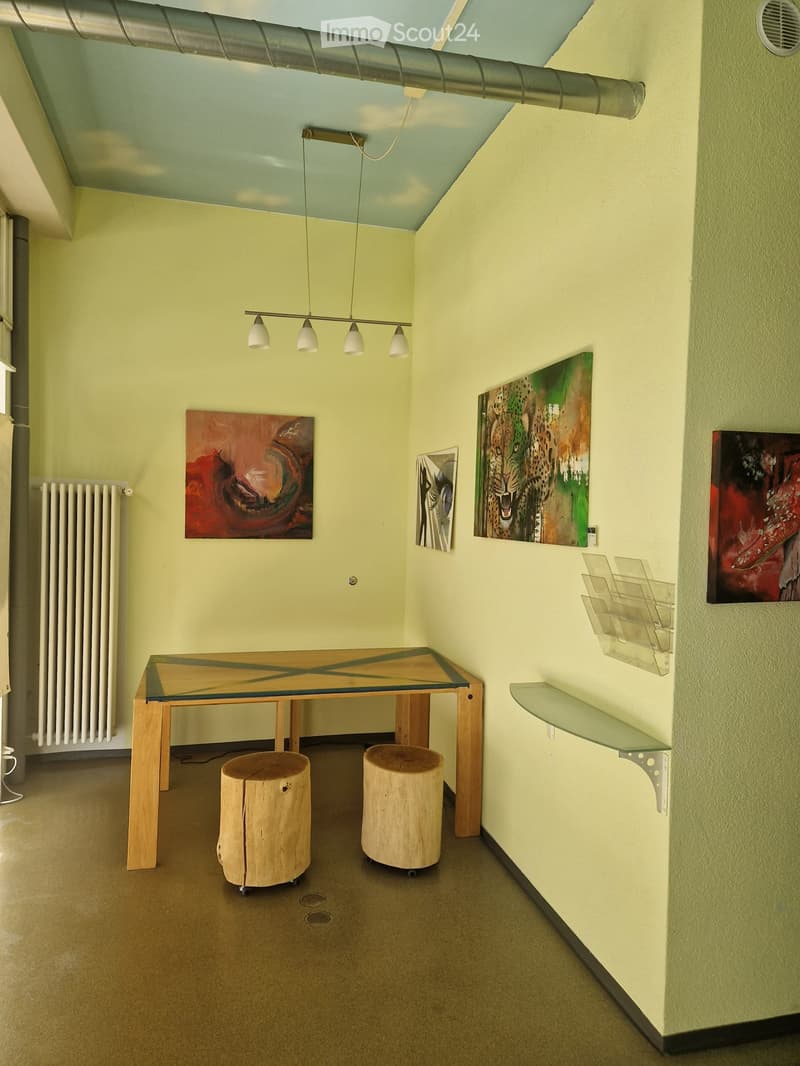 A modern, minimalist room with a glass-topped wooden table, two tree-stump stools, several paintings on the walls, and a glass shelf unit.