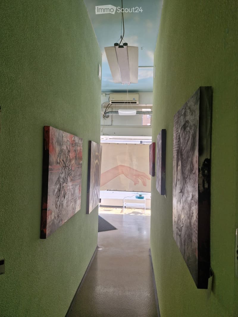 A narrow hallway with green walls, white ceiling, and several large art pieces displayed on the walls. The hallway leads to an open space with a white table and chairs visible in the distance.