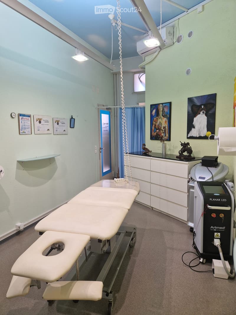 A clean, well-lit medical or treatment room with a treatment table, a medical device labeled "Artmed", a cabinet, framed pictures, and a shelf with decorative items.