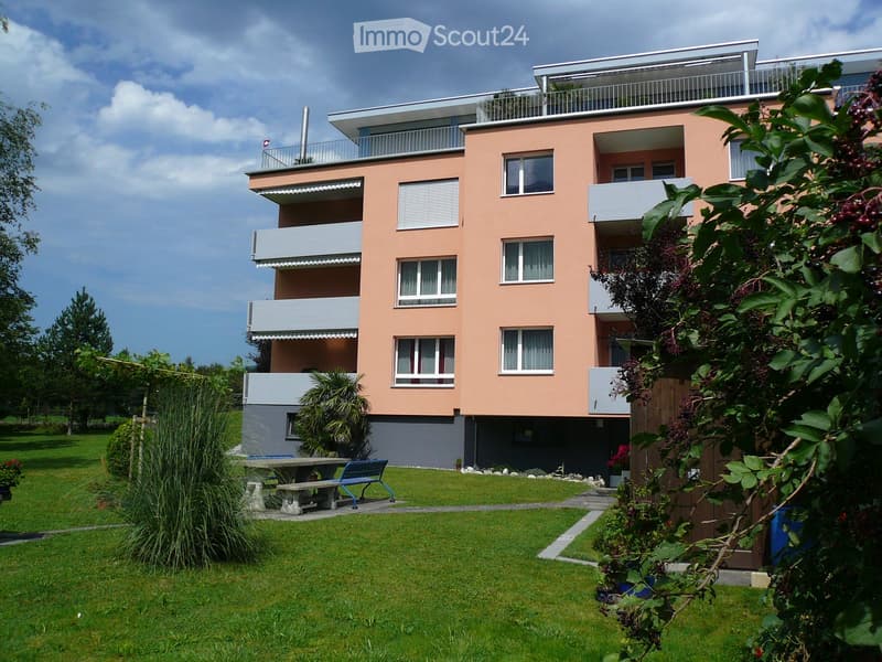 Modern three-story building with pink facade, multiple balconies, and a well-maintained lawn with outdoor seating area.