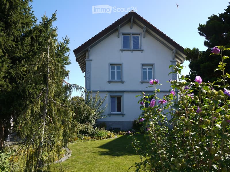 Two-story white house with a red-tiled roof, multiple windows, and a well-maintained garden with green grass and flowering plants.