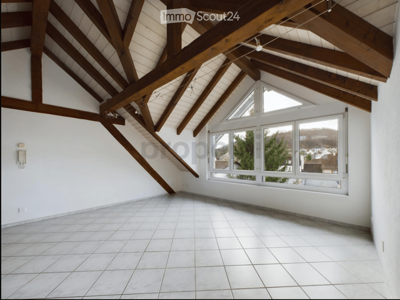 Empty attic room with exposed wooden beams, white tiled floor, large windows offering a view of the surrounding area.