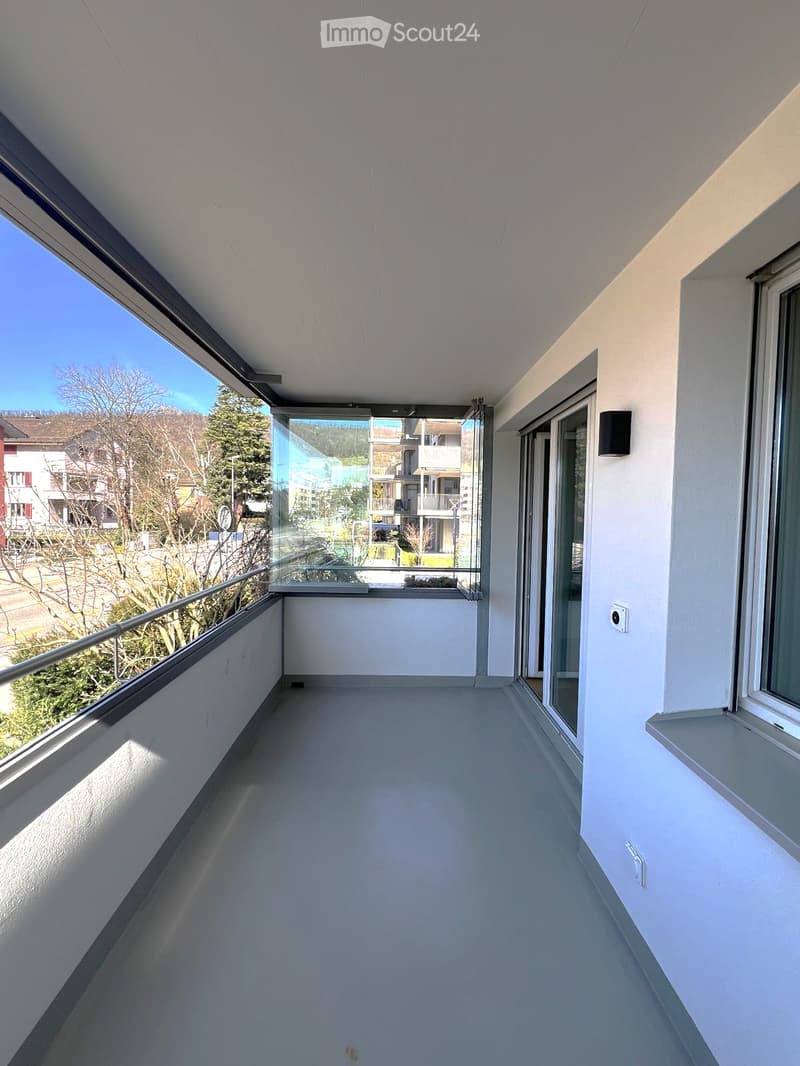 Modern balcony with white walls, large windows, and a metal railing, offering a view of the surrounding area.