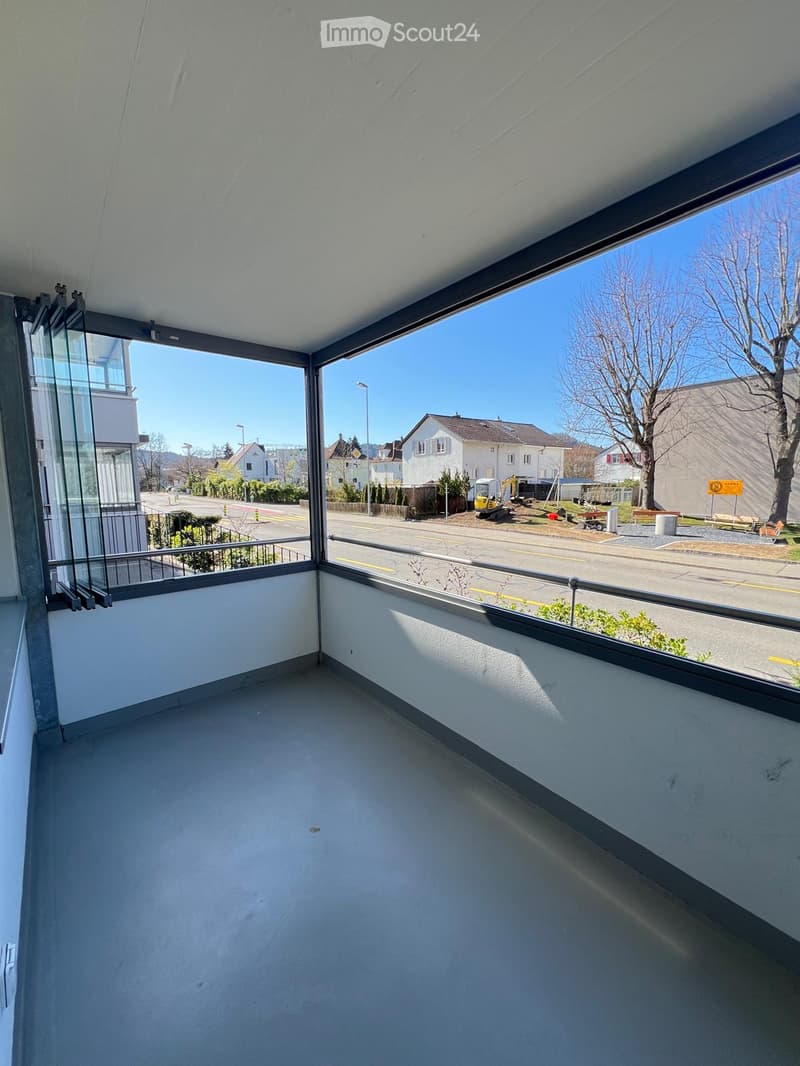 Empty balcony with large windows offering a view of a street, houses, and construction area.