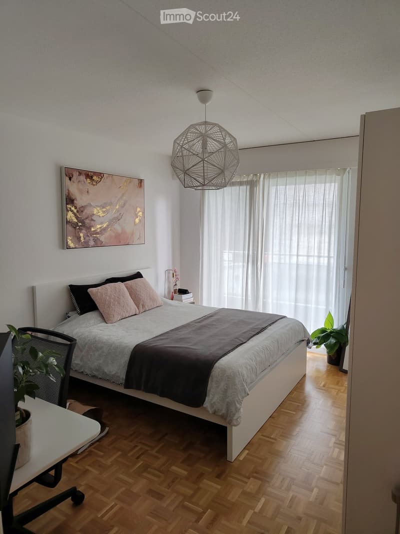 Modern bedroom with a large bed, wooden flooring, and large windows with sheer curtains. Features a geometric pendant light, a small table with a plant, and a painting on the wall.