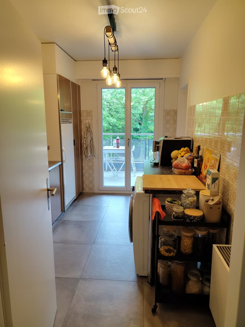 Modern kitchen with white cabinets, stainless steel appliances, and a sliding glass door leading to a balcony. The kitchen features a countertop with various items including fruits, storage jars, and a coffee maker.