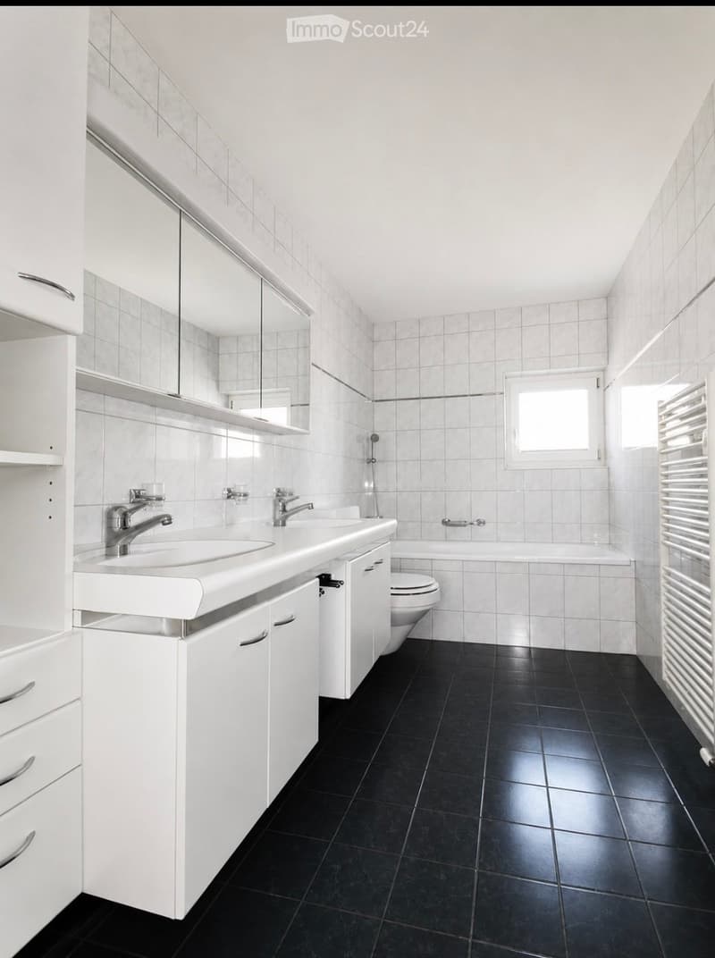 Modern bathroom with white tiles, black floor, double sink, mirror, toilet, bathtub, and radiator.