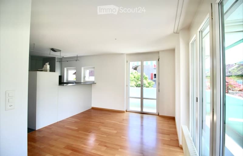 Bright, open-plan living and dining area with wooden flooring, white walls, large glass doors leading to a balcony, and a small kitchen area with a countertop and sink.