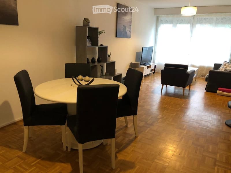 A modern living and dining area with wooden flooring, a round dining table with chairs, a sofa, a television, and a shelf with decorative items.