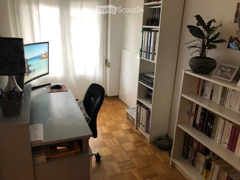 A well-lit study or home office with a wooden floor, a large desk with a computer screen, a chair, a lamp, and shelves filled with books and decorative items.