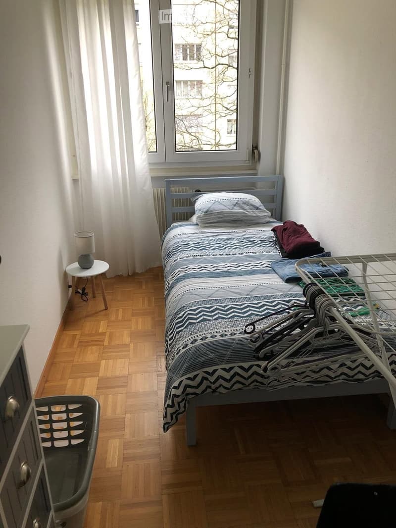 A small bedroom with a single bed, nightstand, lamp, radiator, and a clothing rack. The bed is made with a patterned duvet and pillows, and there are clothes on the bed and rack.