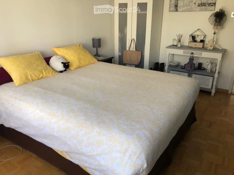 Cozy bedroom with a large bed, yellow pillows, a nightstand with a lamp, a mirror, a dresser with decorative items, and a fan on the wall.