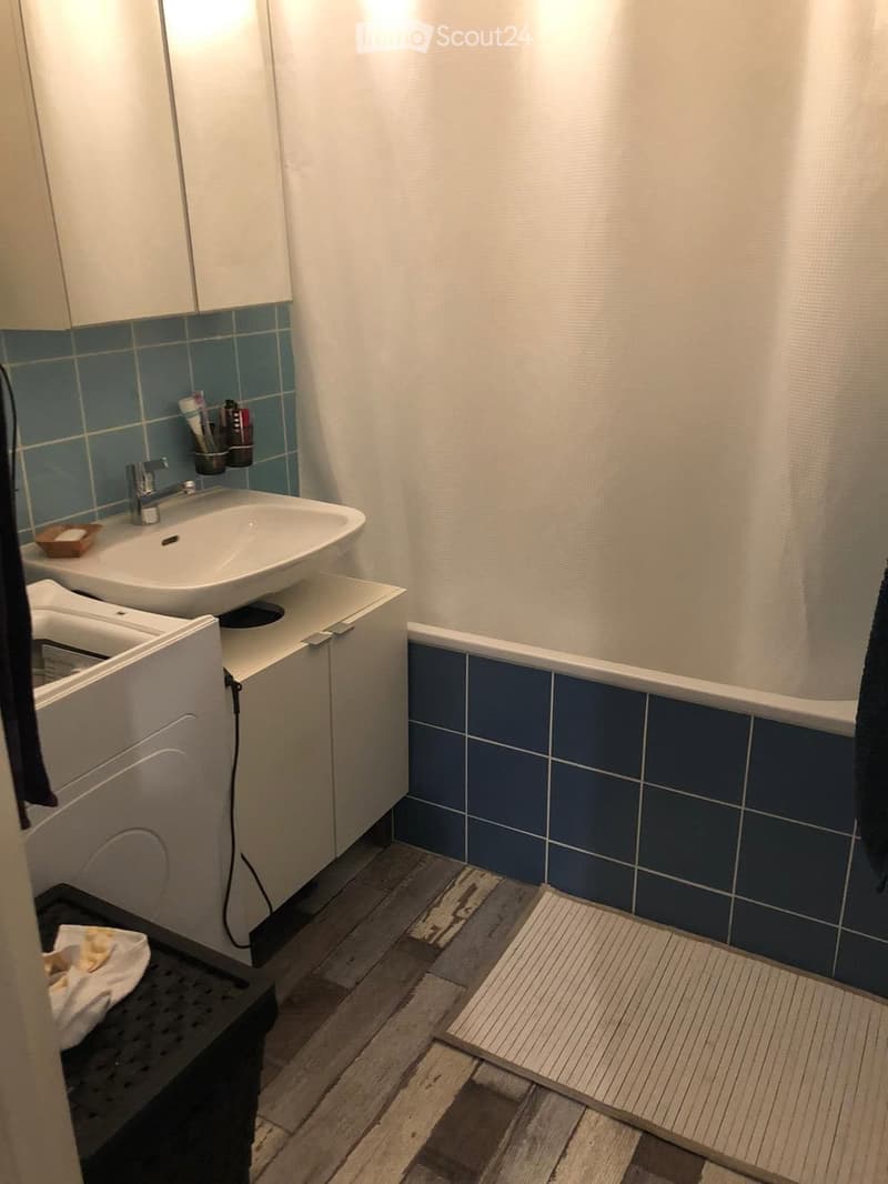 Modern bathroom with a white sink, blue tiles, a bathtub, and a wooden floor. Includes a toothbrush holder, a small cabinet, and a bath mat.