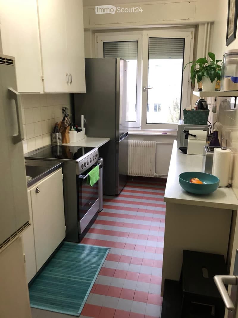 A compact kitchen with white cabinets, stainless steel appliances, and a striped red and white tiled floor. Features include a refrigerator, oven, sink, and various kitchen items on the counter.