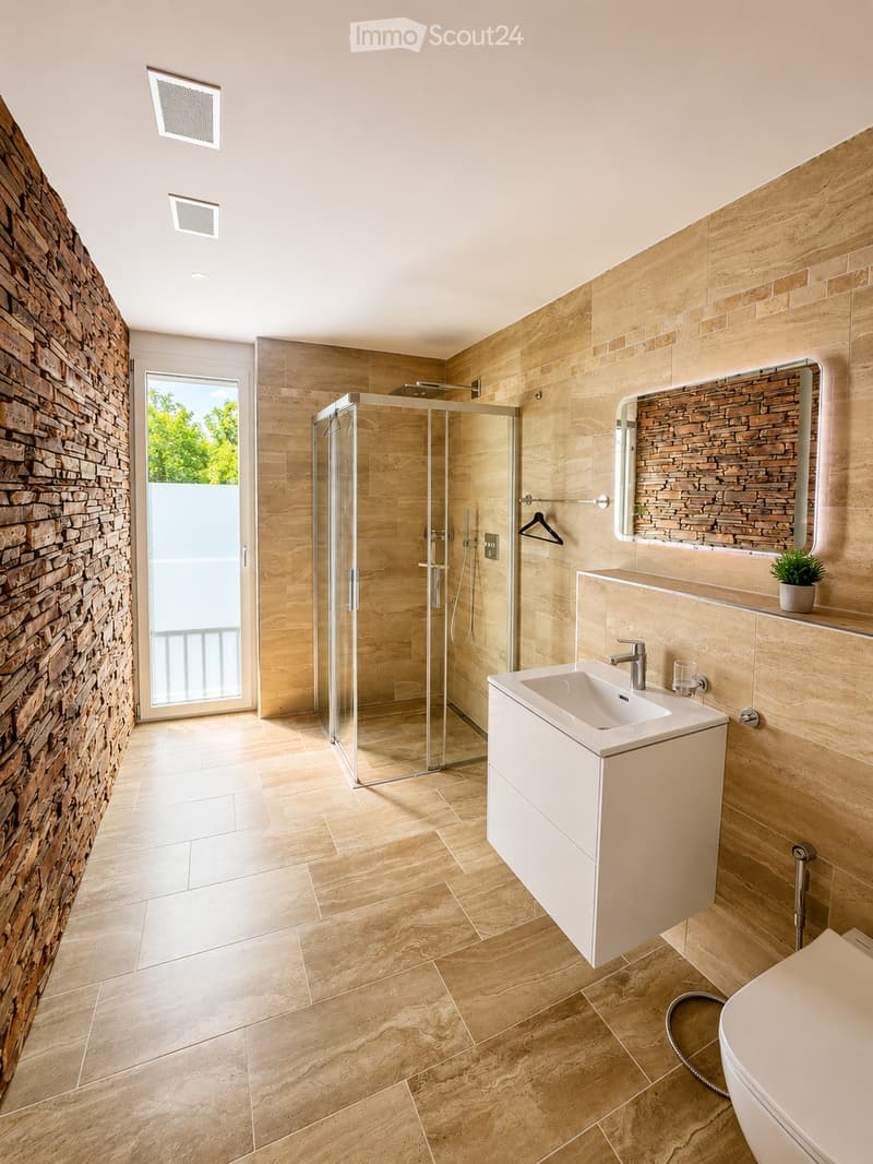 Modern bathroom with beige tiles, a glass-enclosed shower, a white sink, and a toilet. Features a large mirror, a small potted plant, and a view of greenery through a glass door.