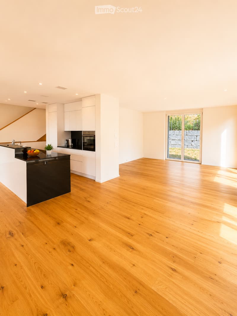 Modern kitchen with white cabinets, black countertops, and a wooden floor. Features a kitchen island, an oven, a faucet, a bowl with fruit, and a small plant.