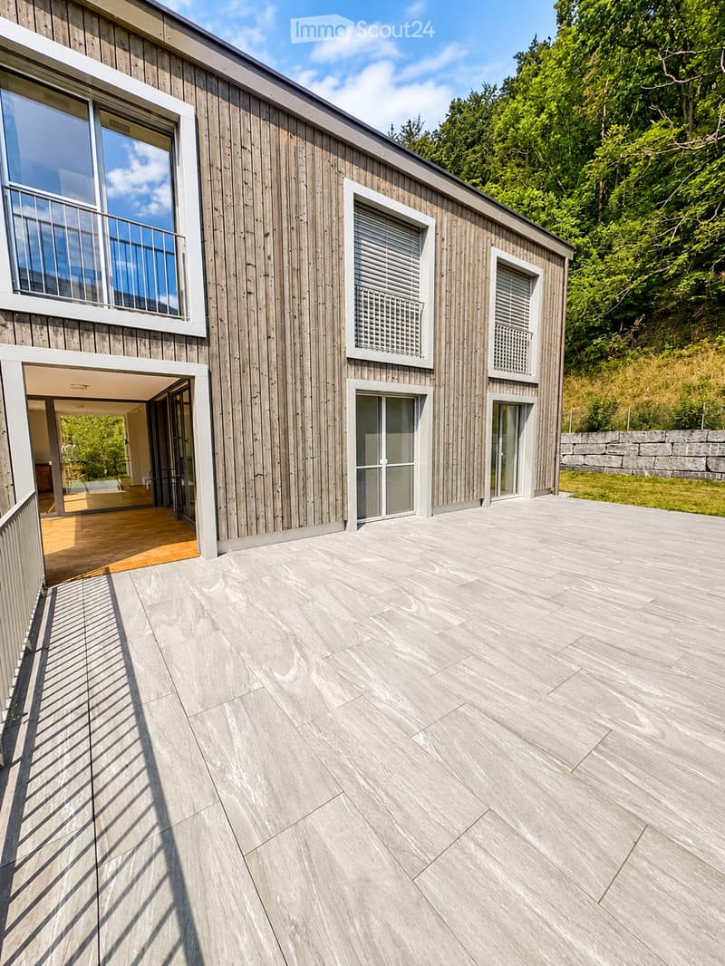 Modern wooden house with large windows, raised ground floor, and a spacious patio area.
