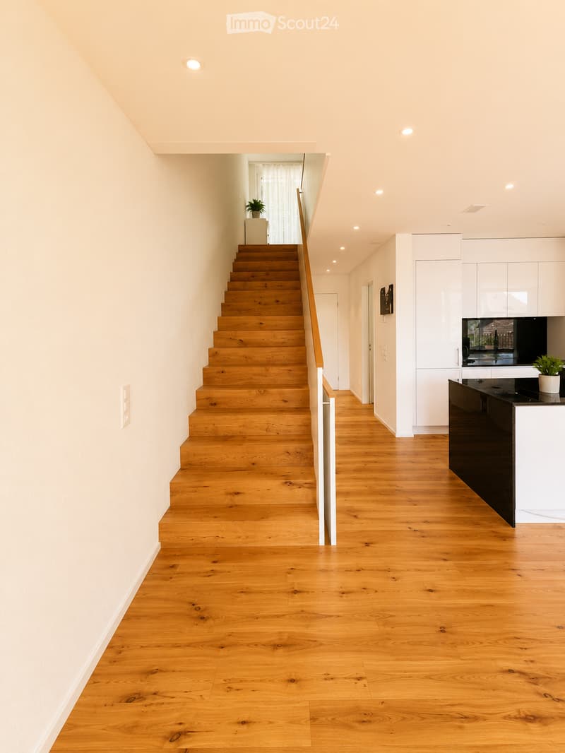 Bright, open hallway with wooden flooring, a staircase leading upwards, and a modern kitchen area visible to the right.