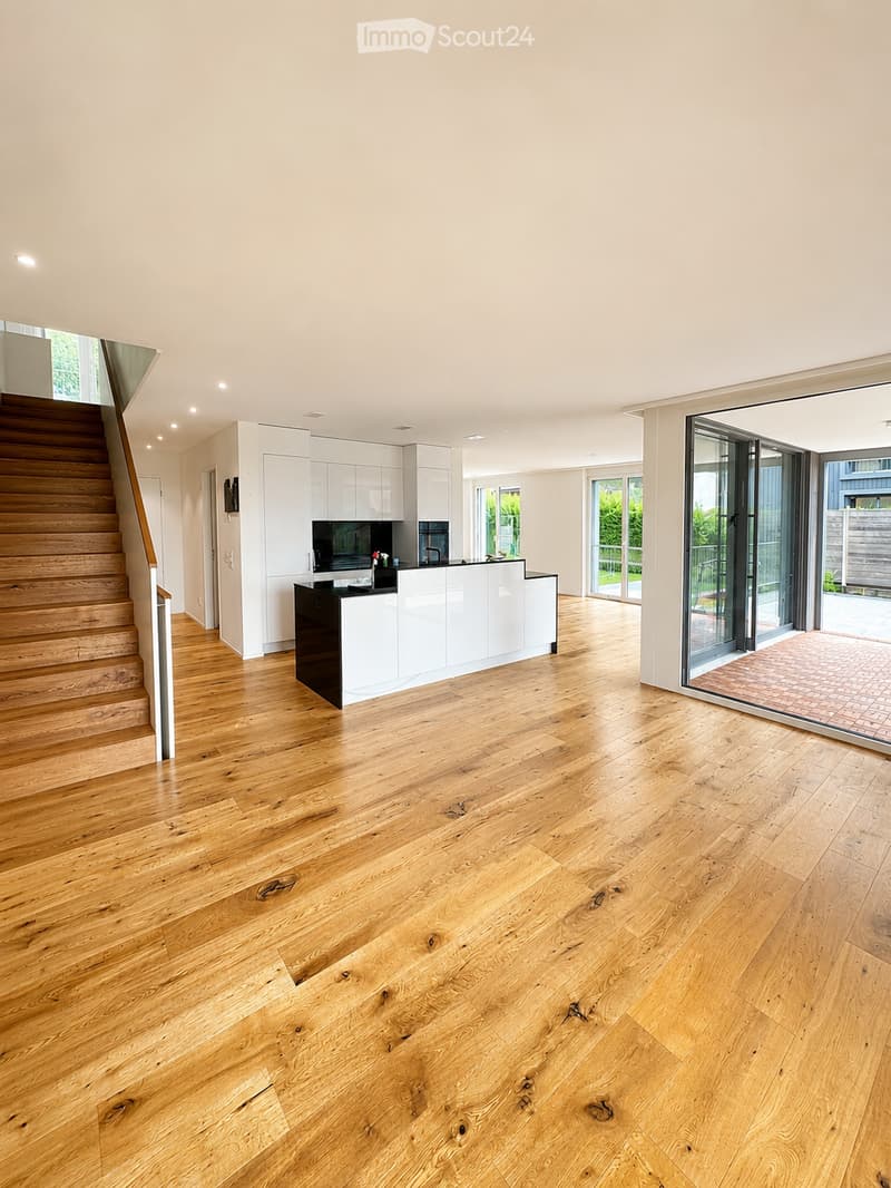 Open-plan kitchen and living area with light wooden flooring, white cabinetry, and a central island. Features a wooden staircase leading to an upper level and large glass doors opening to an outdoor patio.