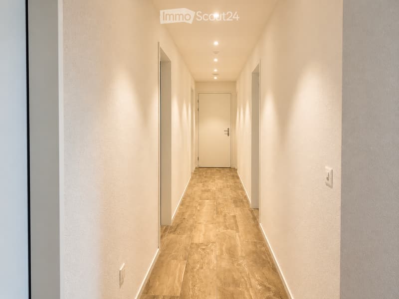 A modern hallway with beige walls, tiled floor, and recessed lighting. Features a closed door at the end and light switches on both sides.