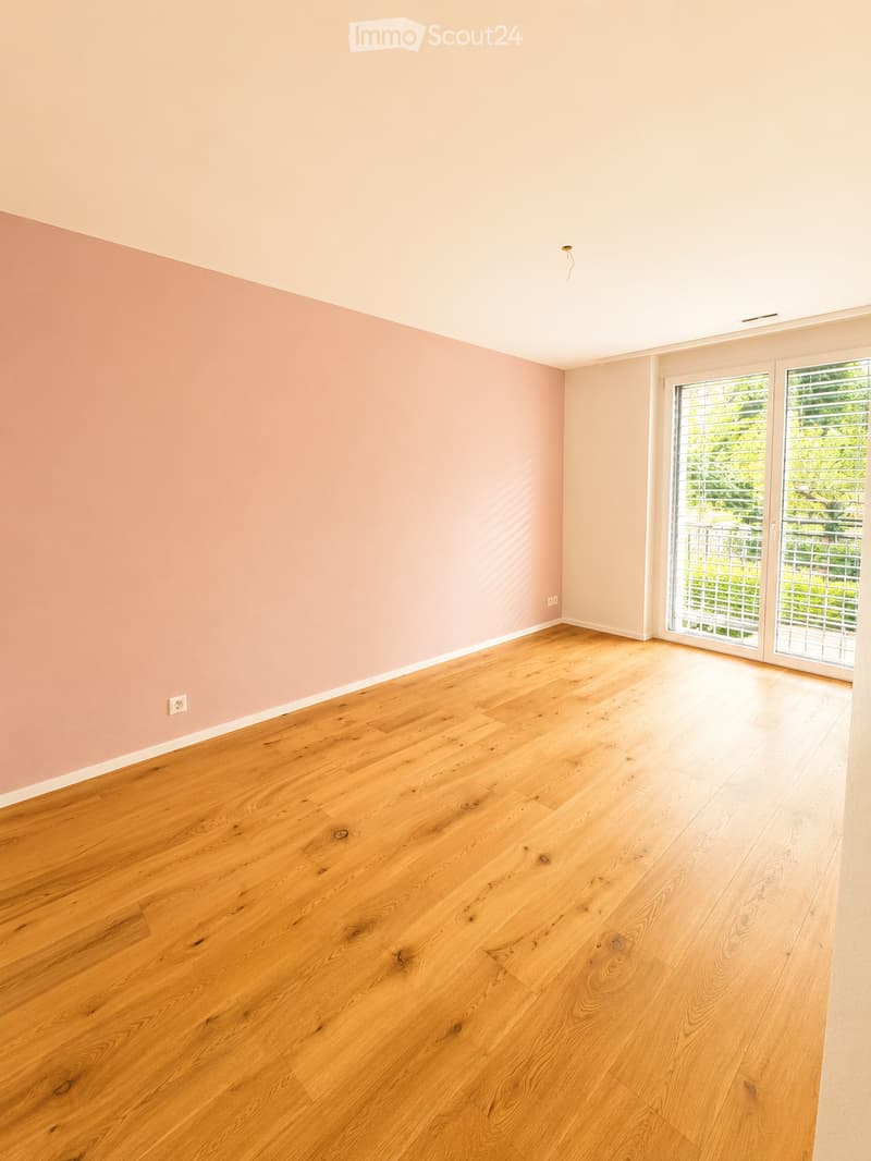 Empty room with light pink walls and polished wooden flooring, featuring large glass doors leading to an outdoor area.