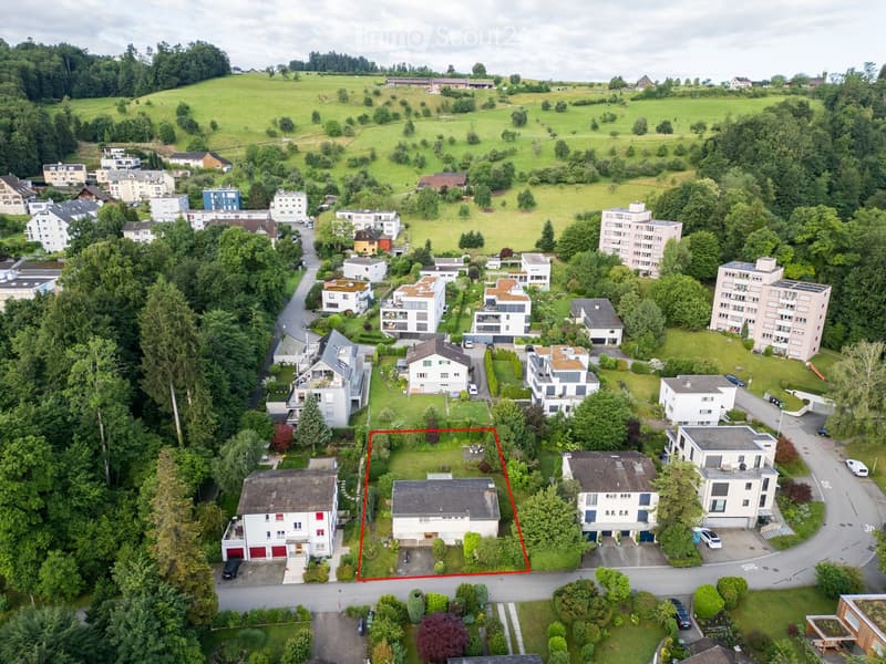 Aerial view of a residential area with multiple houses, green spaces, and trees. The highlighted house is a two-story structure with a garage and a well-maintained garden.