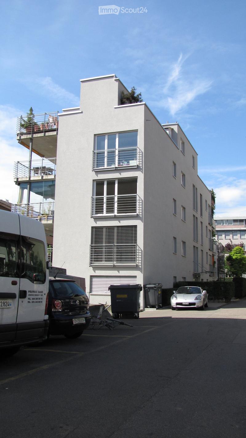 Modern white building with multiple balconies and large windows, surrounded by parked vehicles and greenery.