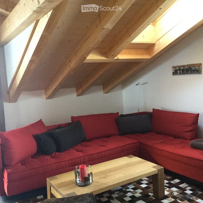 A cozy living room with a red sectional sofa, wooden coffee table, and a candle in a glass holder. The room features exposed wooden beams and a slanted ceiling with skylights.