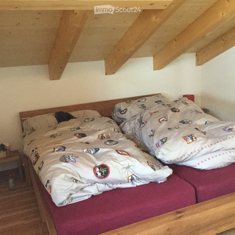 A small bedroom with a wooden bed frame, red mattress, and a duvet with a space-themed design. The room features exposed wooden beams on the ceiling and a nightstand to the left of the bed.