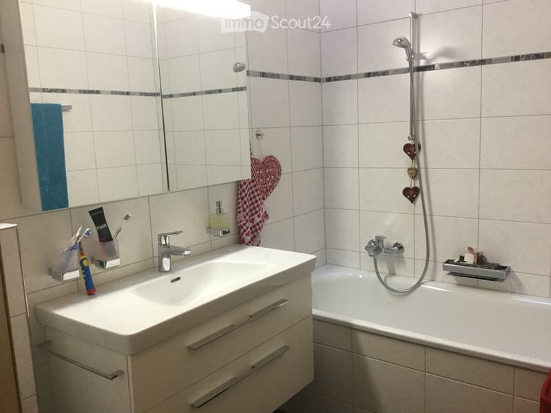 Modern bathroom with a white sink, mirror, toothbrush holder, bathtub, and showerhead. Features a clean, tiled design with decorative heart-shaped ornaments.