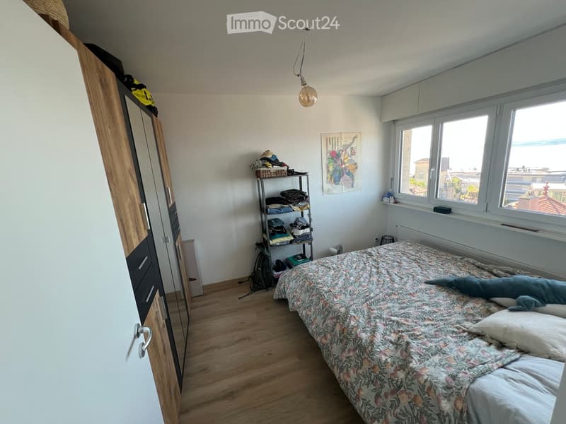 Bedroom with wooden floor, large window, bed with floral bedding, wardrobe, shelf with folded clothes, and a painting on the wall.