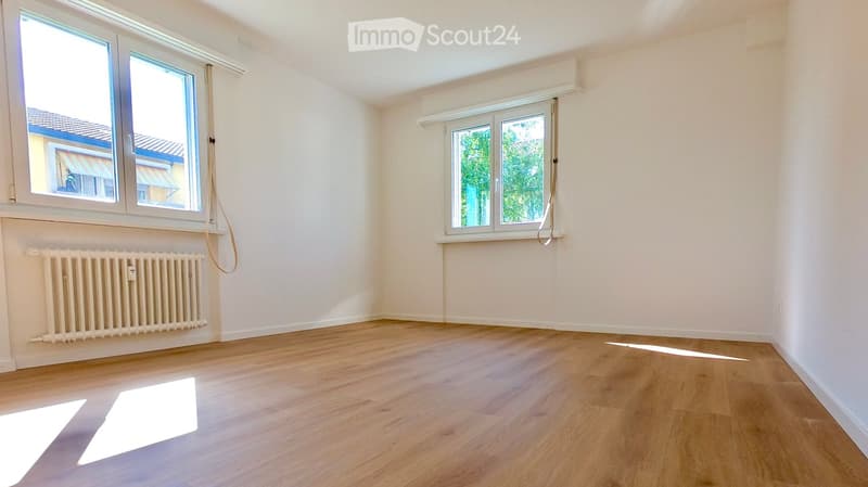 Empty room with wooden flooring, white walls, and large windows allowing natural light to enter. A radiator is visible under one window.