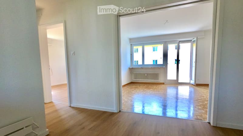 Empty room with wooden flooring, white walls, and a large window with a radiator below it. The room is connected to another empty space through an open doorway.