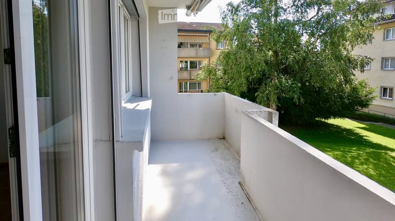 Empty balcony with white walls, concrete floor, and a view of a green lawn and neighboring buildings.