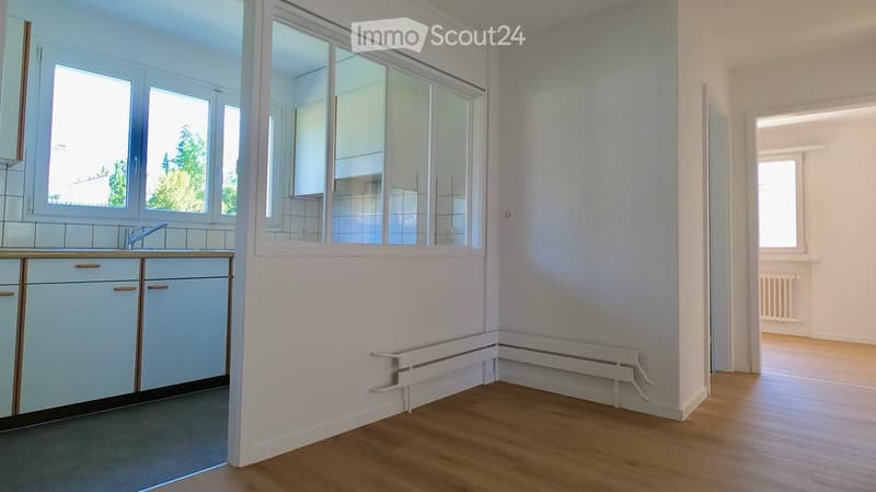 Empty room with wooden flooring, white walls, and built-in cabinets. Large windows provide natural light, and a radiator is visible along the wall.