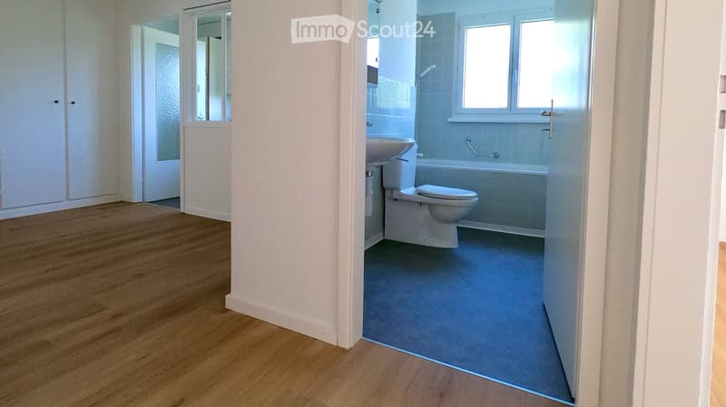 Modern bathroom with white tiles, a toilet, a bathtub, and a sink. The adjacent room has wooden flooring and white walls.