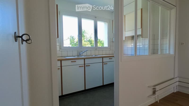 Empty kitchen with white cabinets, a sink, and tiled walls. The room has large windows and a radiator under the windows.