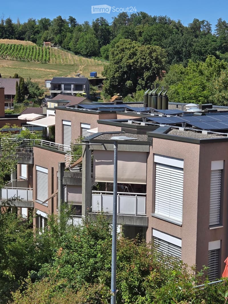 Modern residential building with solar panels on the roof, multiple balconies, and surrounding greenery.