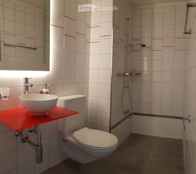 Modern bathroom with white tiles, red accent shelf, sink, toilet, and shower area.