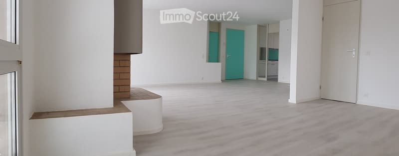 Empty room with white walls, grey carpeted floor, and a visible kitchen area with cabinets and a countertop.