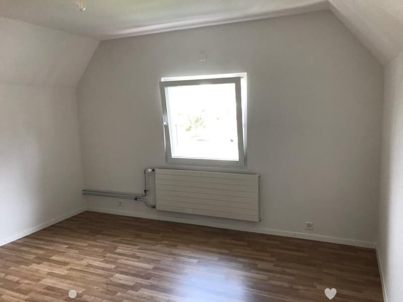 Empty attic room with wooden flooring, white walls, a single window, and a radiator. No furniture or personal items present.