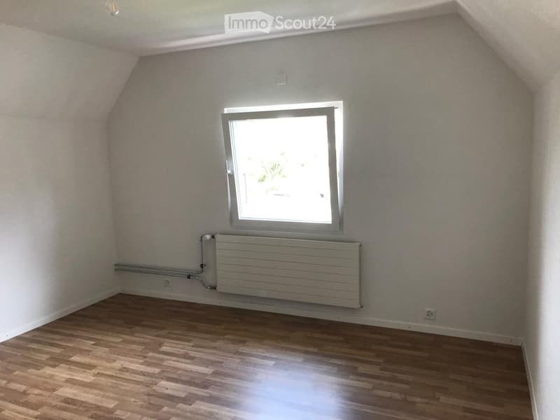 Empty attic room with wooden flooring, white walls, a single window, and a radiator. No furniture or personal items present.