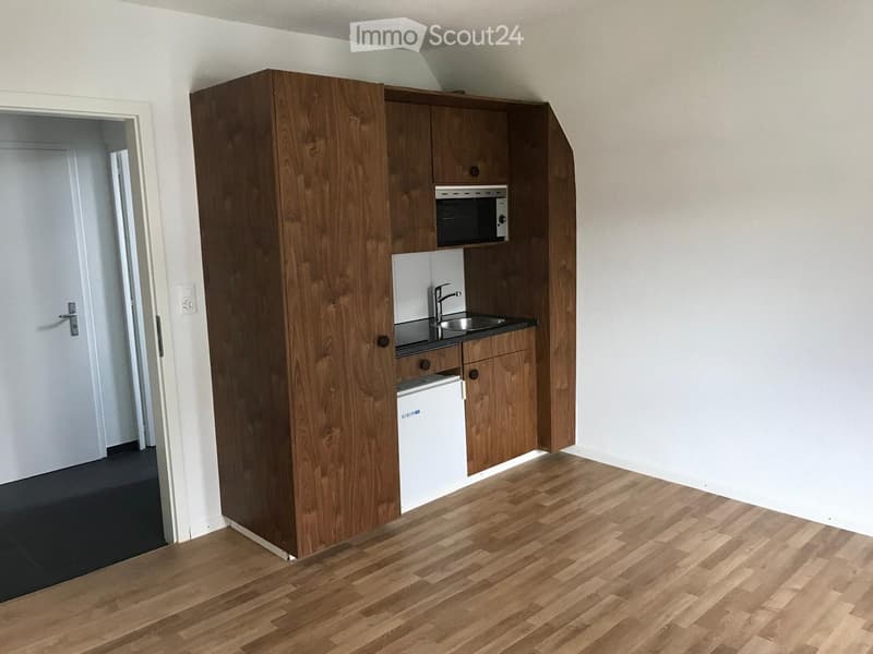 Compact kitchen with wooden cabinets, a sink, microwave, and refrigerator, featuring light wooden flooring and white walls.