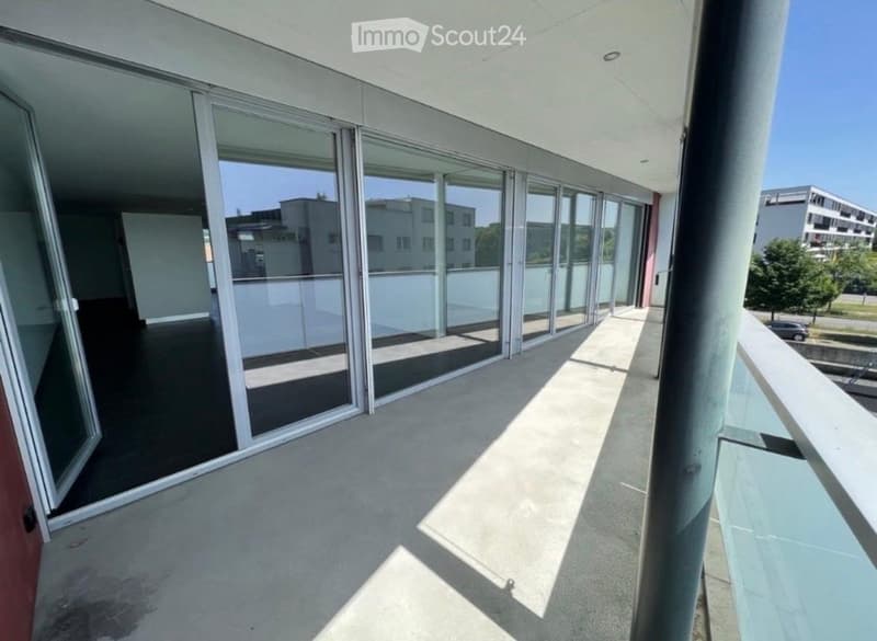 Modern balcony with large glass sliding doors, concrete floor, and metal railing, offering a view of surrounding buildings and greenery.