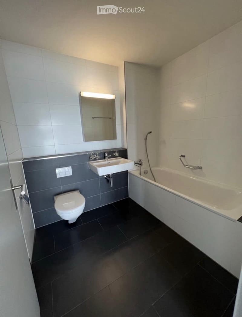 Modern bathroom with a toilet, sink, bathtub, and mirror. Features dark tiled flooring and grey tiled walls.