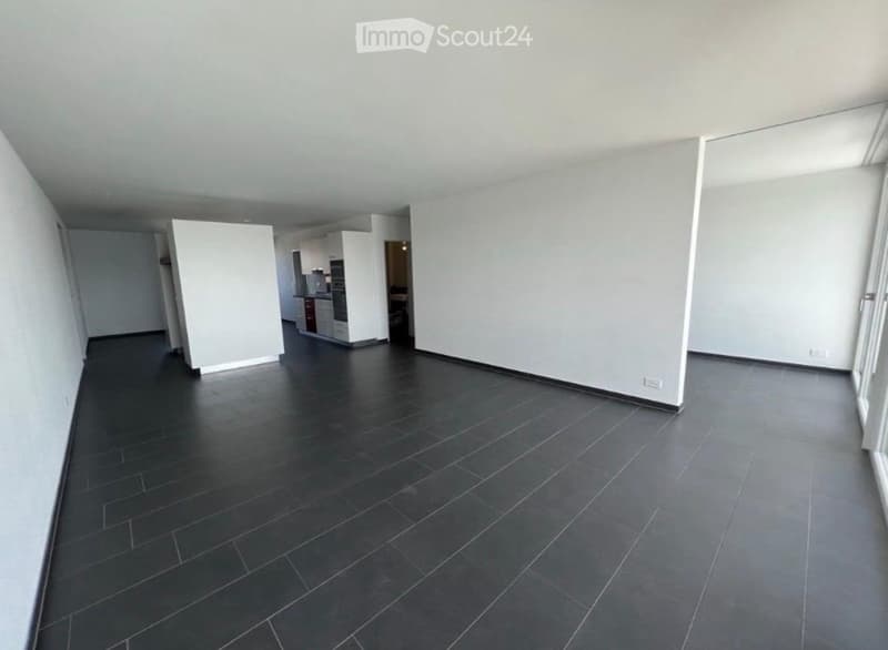 Empty room with dark tiled floor and white walls, featuring an open doorway leading to a kitchen area with white cabinets and appliances.