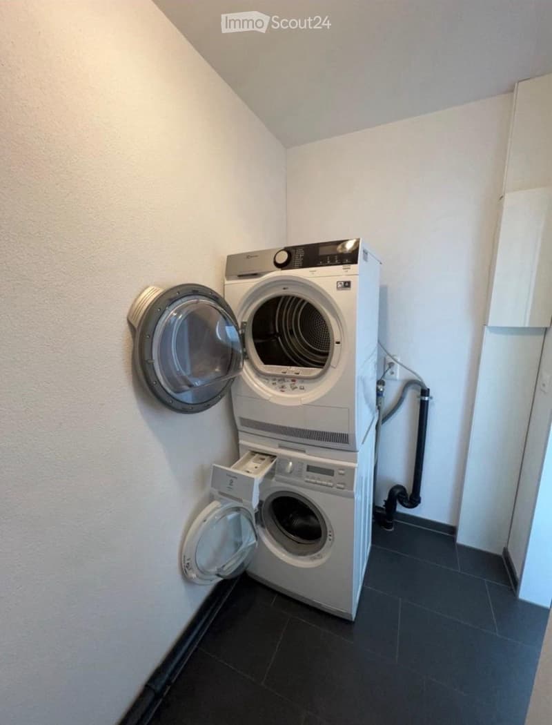 Compact bathroom with a stacked washing machine and dryer unit, both with open doors, against a white wall with a dark tiled floor.