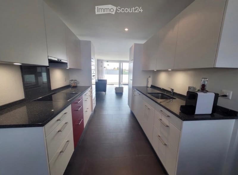 Modern kitchen with white cabinets, black granite countertops, stainless steel sink, and various appliances including a toaster and coffee maker. The space is open and well-lit, with a view to a living area beyond.