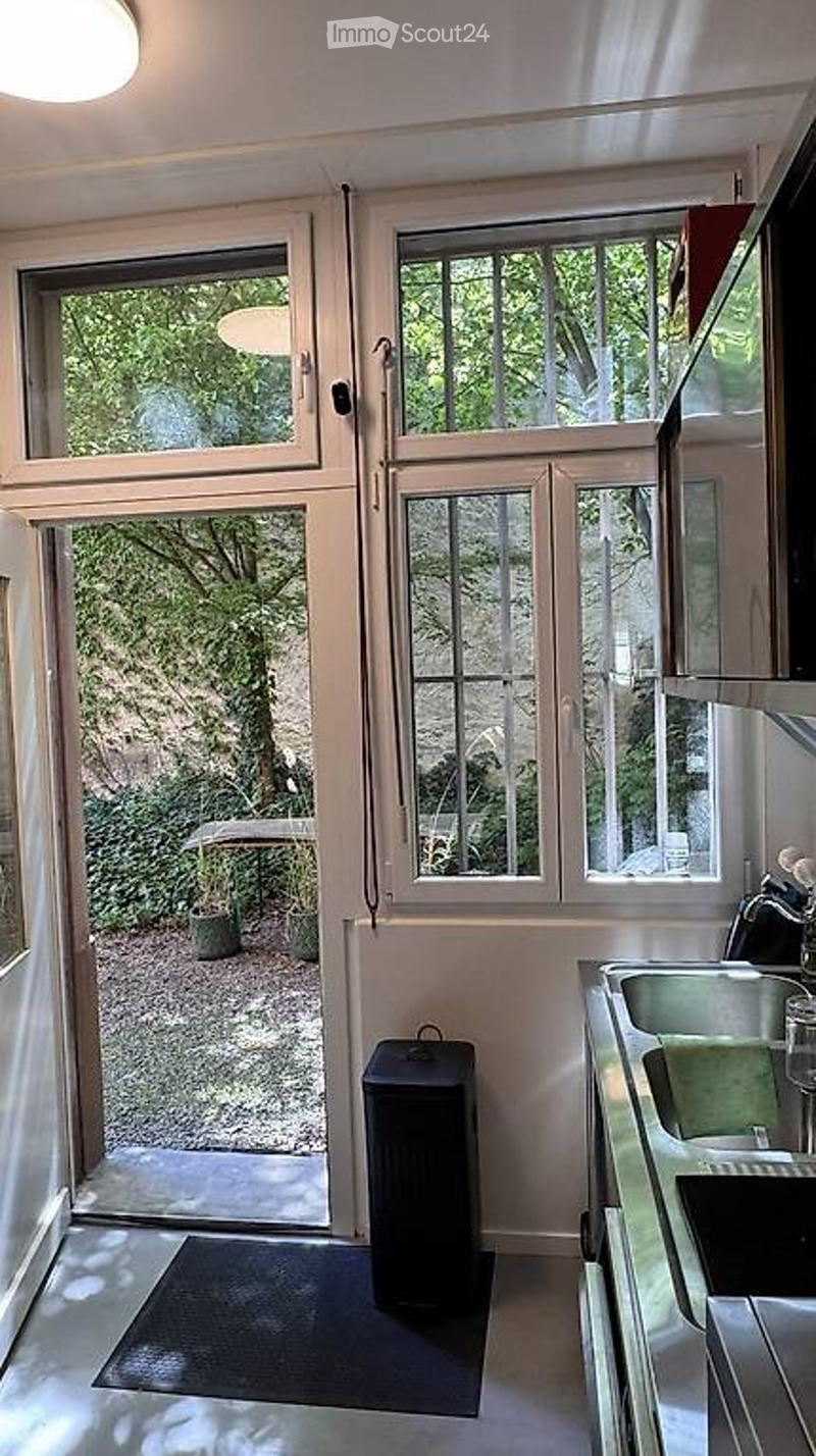 Modern kitchen with stainless steel sink, large windows overlooking a garden, and a black trash bin near the door.